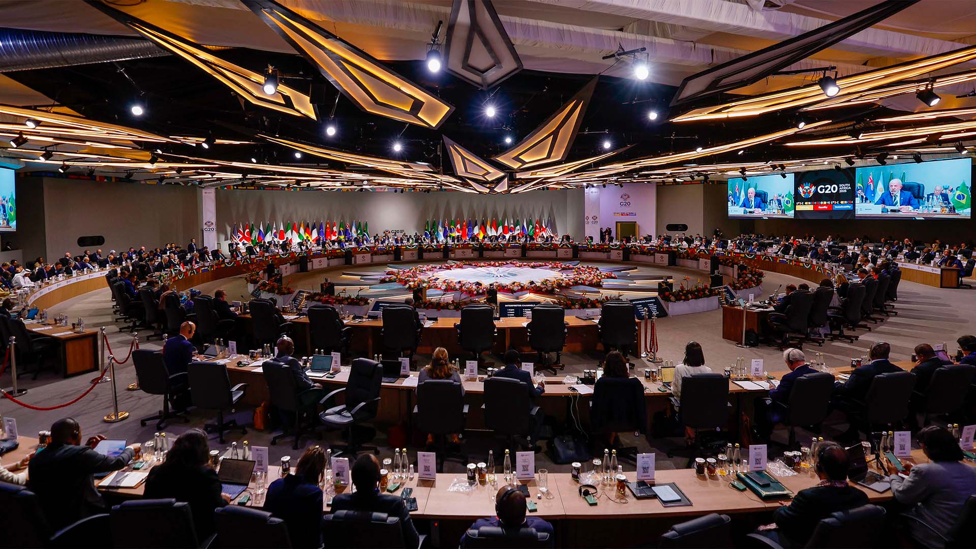 Delegados de los países asistentes a la reunión del G20 en la sala de conferencias del Centro de Exposiciones de Johannesburgo (22/11/2025) en la Cumbre del G20 de 2025 en Sudáfrica. Todos están sentados alrededor de varios círculos concéntricos formados por las propias mesas, y en el centro, un círculo con formas geométricas y flores decora el espacio. En el techo, se iluminan varias formas geométricas y, al fondo, se pueden ver las banderas de los países participantes.