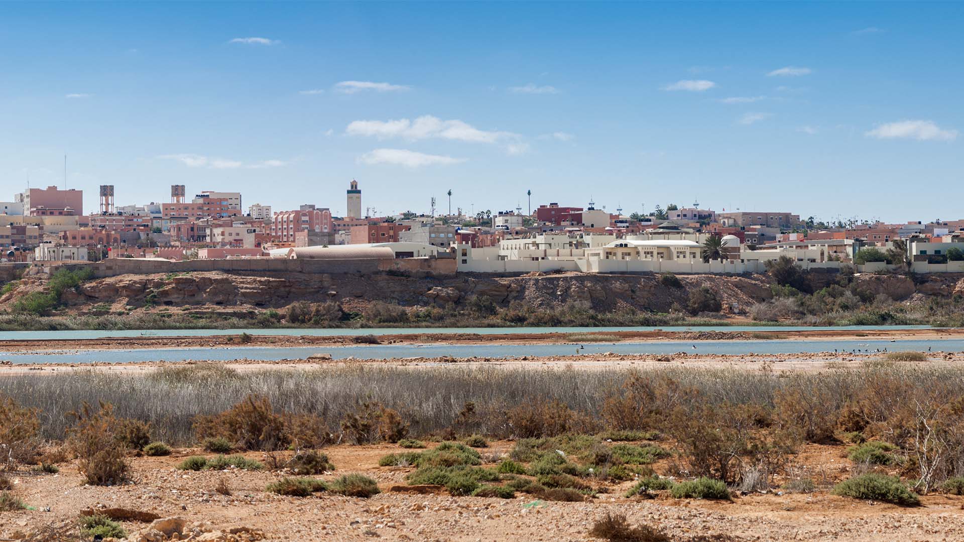 Vista panorámica de la ciudad de El Aaiún, en el territorio del Sáhara Occidental. En primer plano, un terreno árido con vegetación dispersa; más atrás, una superficie extensa de agua; y, al fondo, una vista general de la ciudad de El Aaiún.