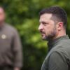 Foto de perfil del presidente de Ucrania, Volodímir Zelenski (cabello corto y barba, con la vestimenta verde militar) sobre un fondo de vegetación desenfocado, y otra persona similar aparece borrosa al fondo. Tomada durante el día de las fuerzas armadas ucranianas en agosto de 2025.