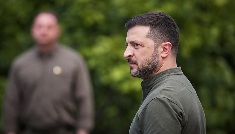Foto de perfil del presidente de Ucrania, Volodímir Zelenski (cabello corto y barba, con la vestimenta verde militar) sobre un fondo de vegetación desenfocado, y otra persona similar aparece borrosa al fondo. Tomada durante el día de las fuerzas armadas ucranianas en agosto de 2025.