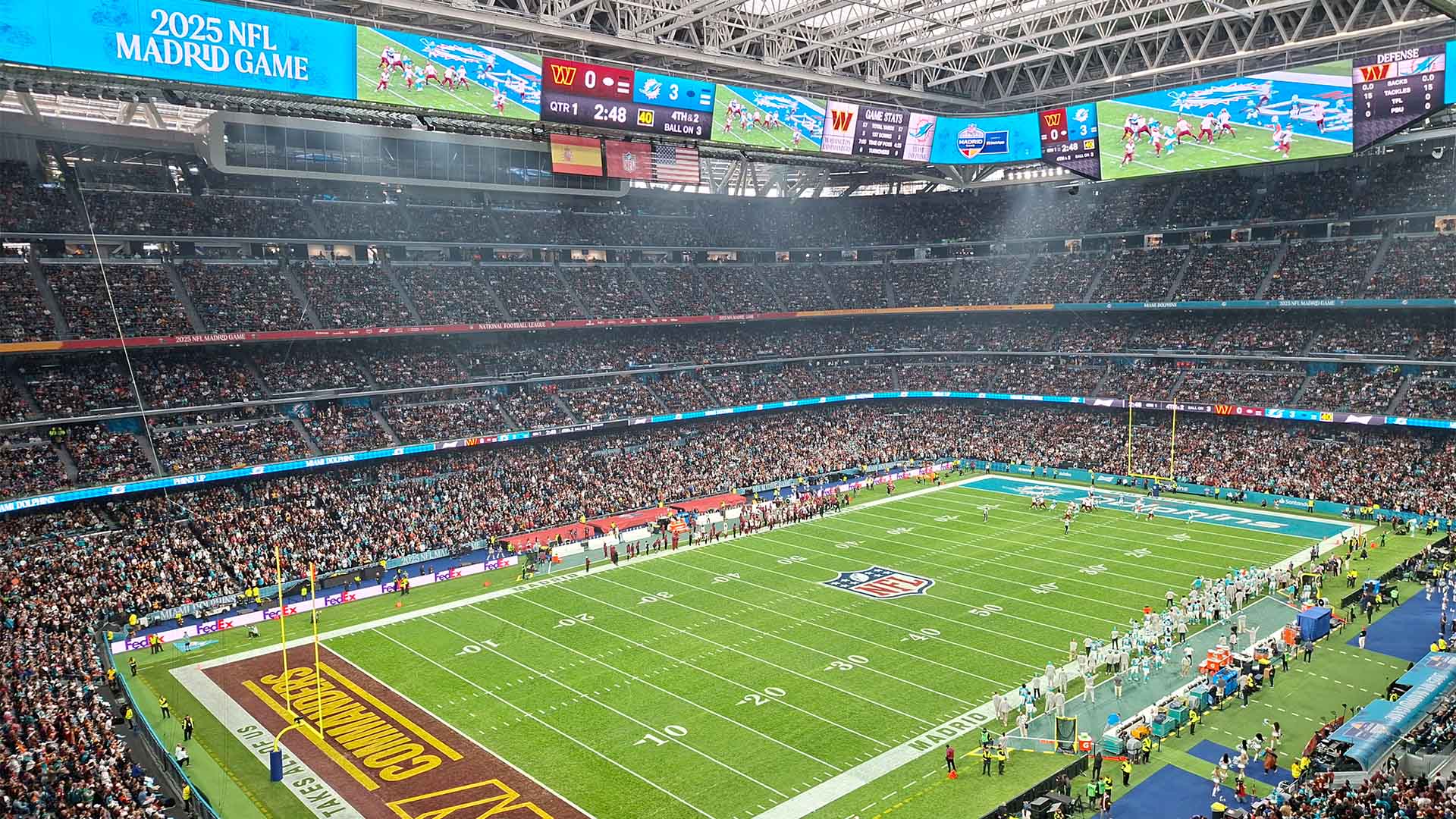 Vista del estadio Santiago Bernabéu durante el partido NFL Miami Dolphins vs Washington Commanders (16/11/2025). El campo está vacío y los jugadores se encuentran agrupados fuera de él, cada equipo a un lado. En el centro del campo, el logo de la NFL y a los lados los logos de ambos equipos. Las gradas están llenas y en la pantalla superior aparece el fondo azul de los Dolphins con el texto “2025 NFL Madrid Game”, la retransmisión en directo y el marcador: Commanders 0 – Dolphins 3. Sobre la pantalla cuelgan las banderas de España, la NFL y Estados Unidos.