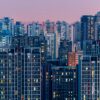 Vista panorámica al atardecer de una zona urbana densamente poblada en Pekín, con numerosos edificios residenciales que se extienden en diferentes planos. Algunas ventanas iluminadas contrastan con la luz cálida del ocaso, mientras al fondo un cielo rosado anuncia el final del día.