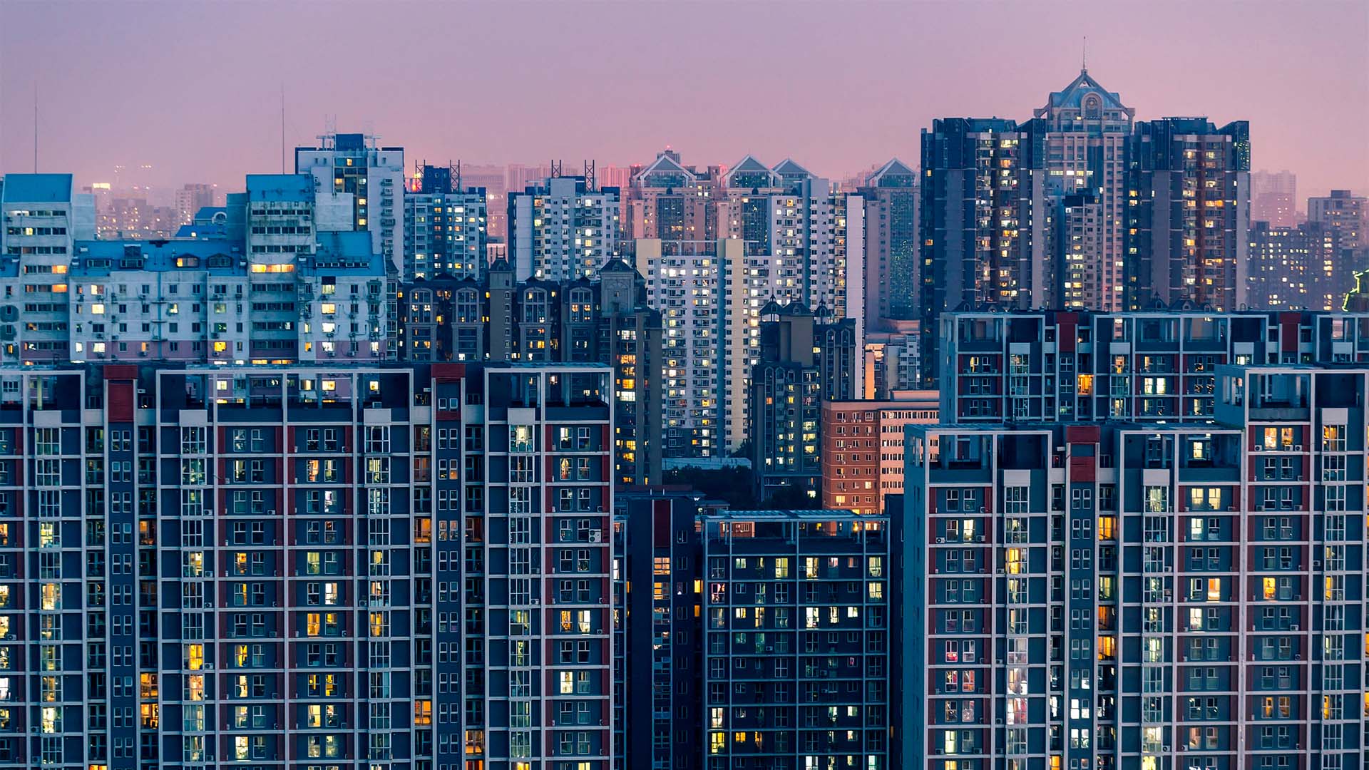 Vista panorámica al atardecer de una zona urbana densamente poblada en Pekín, con numerosos edificios residenciales que se extienden en diferentes planos. Algunas ventanas iluminadas contrastan con la luz cálida del ocaso, mientras al fondo un cielo rosado anuncia el final del día.