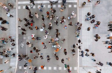Vista superior de un cruce peatonal muy concurrido en España. A ambos lados, dos aceras amplias donde los transeúntes esperan para cruzar en el semáforo. En el centro, el paso de peatones está abarrotado de personas que avanzan en ambas direcciones. En la parte superior, hay vehículos detenidos antes del semáforo, mientras que en la inferior aparece parte de los semáforos que regulan el cruce.