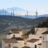 En primer plano, un conjunto de casas de fachada blanca y cubierta a dos aguas de ladrillo marrón del pueblo de Casares, Málaga. Detrás, un paisaje verde, con un parque de turbinas eólicas al fondo, y una sombra montañosa en el horizonte.
