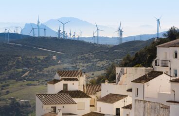 En primer plano, un conjunto de casas de fachada blanca y cubierta a dos aguas de ladrillo marrón del pueblo de Casares, Málaga. Detrás, un paisaje verde, con un parque de turbinas eólicas al fondo, y una sombra montañosa en el horizonte.