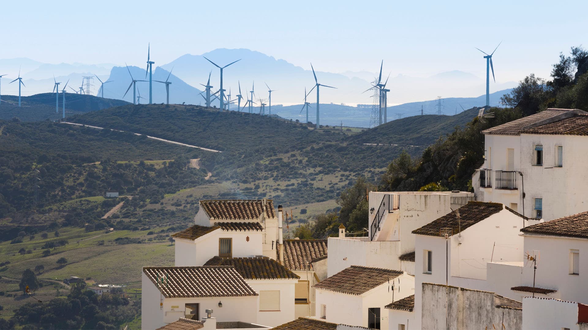 En primer plano, un conjunto de casas de fachada blanca y cubierta a dos aguas de ladrillo marrón del pueblo de Casares, Málaga. Detrás, un paisaje verde, con un parque de turbinas eólicas al fondo, y una sombra montañosa en el horizonte.