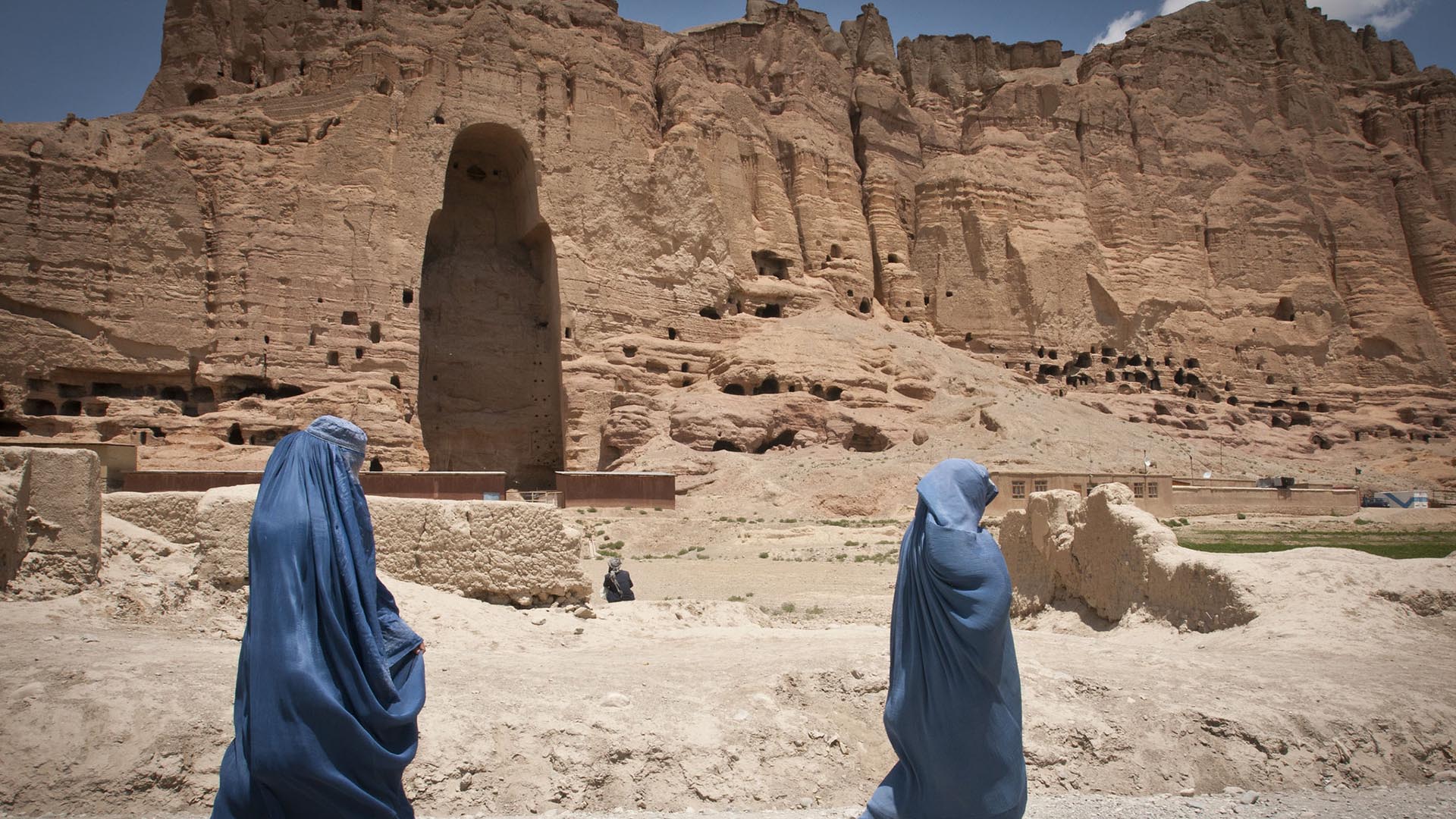 Dos mujeres caminan cubiertas con burkas de color azul en Bamiyán, provincia de Afganistán en 2012. Al fondo del paisaje árido, una cadena de montañas con una cavidad vacía donde se erguía uno de los Budas de Bamiyán construidos en los años 507 y 554 d.C. y destruidos por los talibanes en 2001.