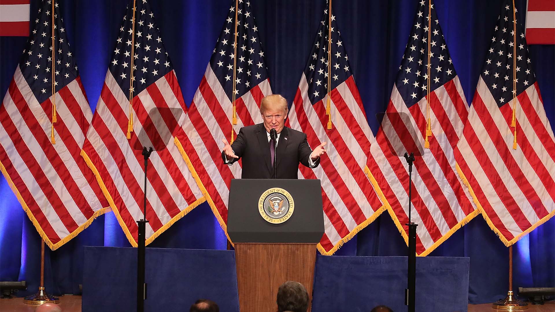 Donald Trump, presidente de EEUU, presenta la Estrategia de Seguridad Nacional de 2017 en el Ronald Reagan Building de Washington D.C. (18/12/2017). Trump, con gesto inquisitivo, está en el centro del del escenario tras un atril con el sello presidencial. Tras él, seis banderas estadounidenses y, al fondo, un telón azul oscuro. En 2025, no ha habido una presentación específica para el documento.