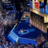 El público rodea el escenario, entre el que hay banderas de EEUU y del Partido Demócrata, así como otras con los nombres de Harris y Walz. Kamala Harris está en el centro del escenario. A ambos lados, hay unas tribunas con más representantes de los demócratas. Detrás del escenario se proyectan banderas de EEUU y varias pantallas que muestran el texto “DNC 2024”, siglas de Democratic National Convention.