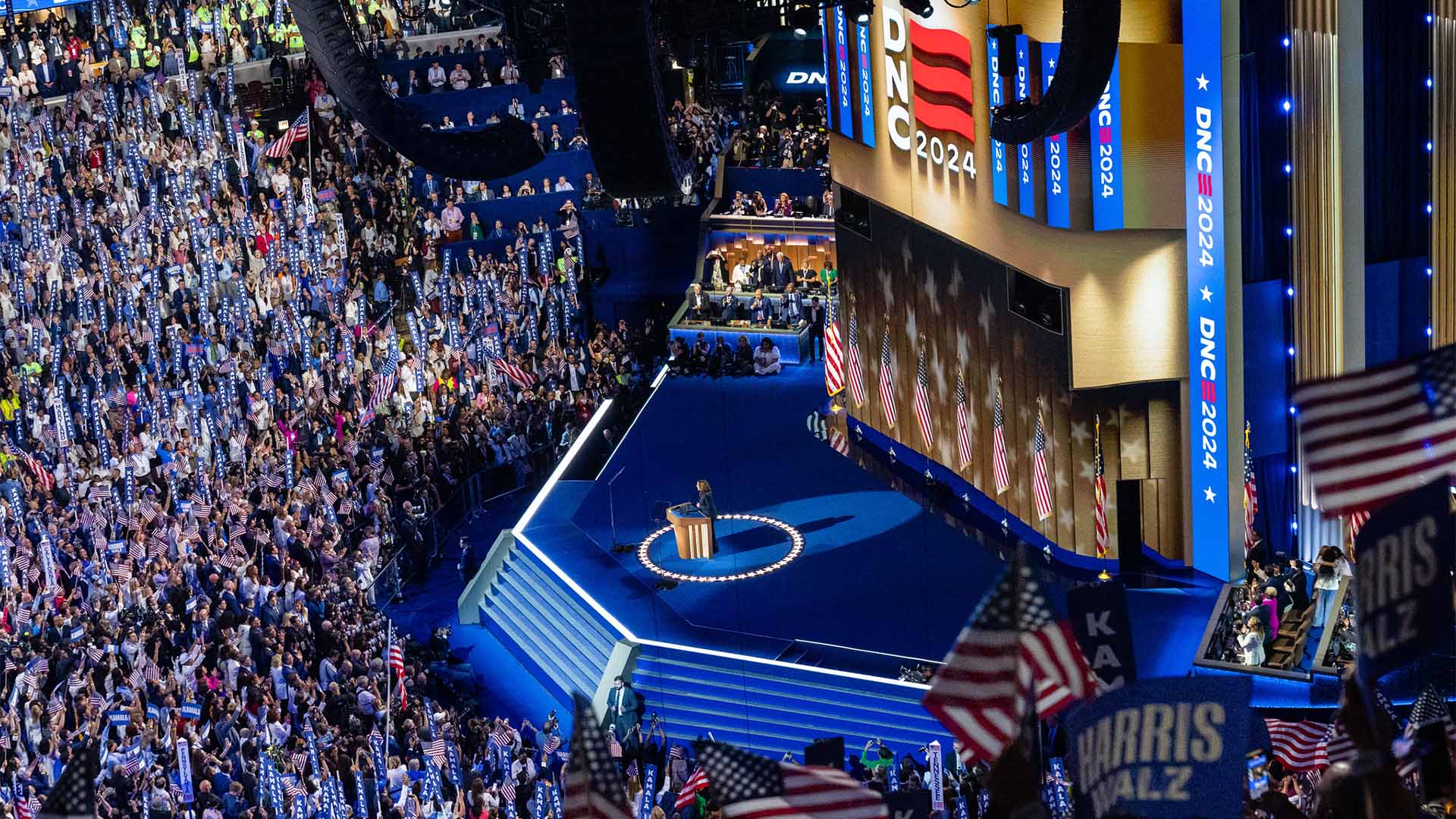 El público rodea el escenario, entre el que hay banderas de EEUU y del Partido Demócrata, así como otras con los nombres de Harris y Walz. Kamala Harris está en el centro del escenario. A ambos lados, hay unas tribunas con más representantes de los demócratas. Detrás del escenario se proyectan banderas de EEUU y varias pantallas que muestran el texto “DNC 2024”, siglas de Democratic National Convention.
