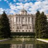 The north façade of the Royal Palace of Madrid rises imposingly with its characteristic classical Baroque architecture, built in the 18th century, with white and grey tones that stand out against a cloudy blue sky. The perspective of the building is taken from one of the fountains in the Sabatini Gardens, surrounded by sculptures and various hedges. Three tall trees stand out, creating a visual bridge between the garden and the palace.