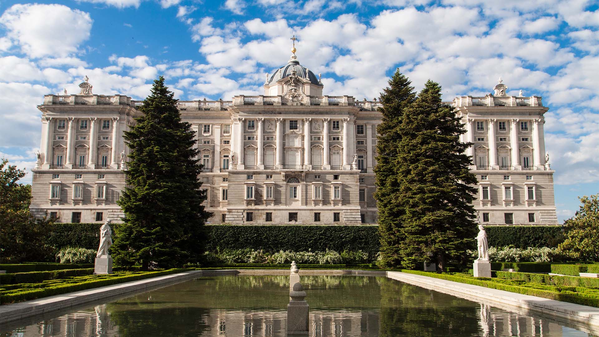 The north façade of the Royal Palace of Madrid rises imposingly with its characteristic classical Baroque architecture, built in the 18th century, with white and grey tones that stand out against a cloudy blue sky. The perspective of the building is taken from one of the fountains in the Sabatini Gardens, surrounded by sculptures and various hedges. Three tall trees stand out, creating a visual bridge between the garden and the palace.