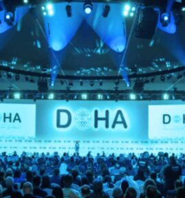 Large conference room at the Doha Forum with attendees in front of a blue-lit stage and two screens displaying the word ‘DOHA’.