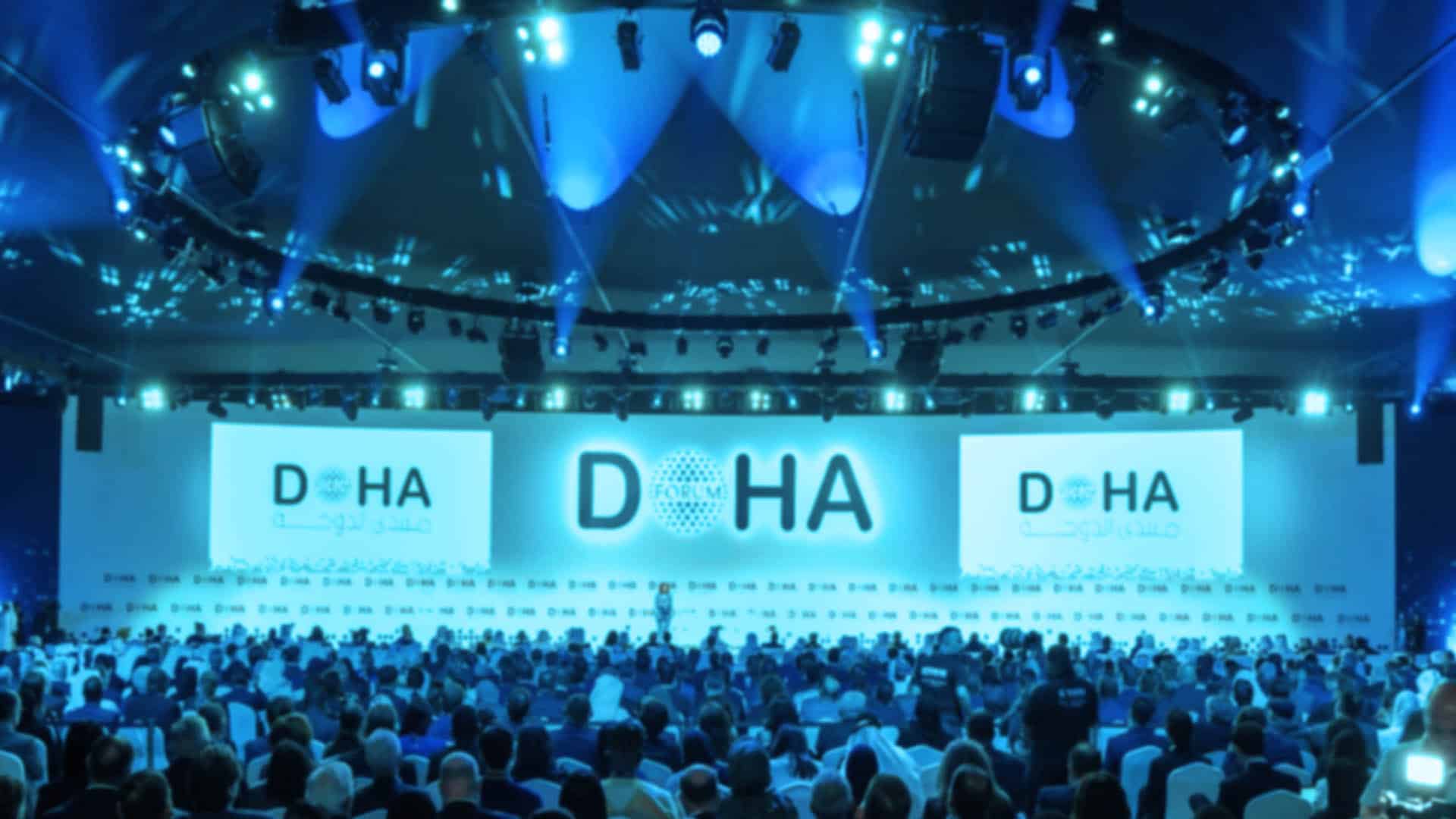 Large conference room at the Doha Forum with attendees in front of a blue-lit stage and two screens displaying the word ‘DOHA’.