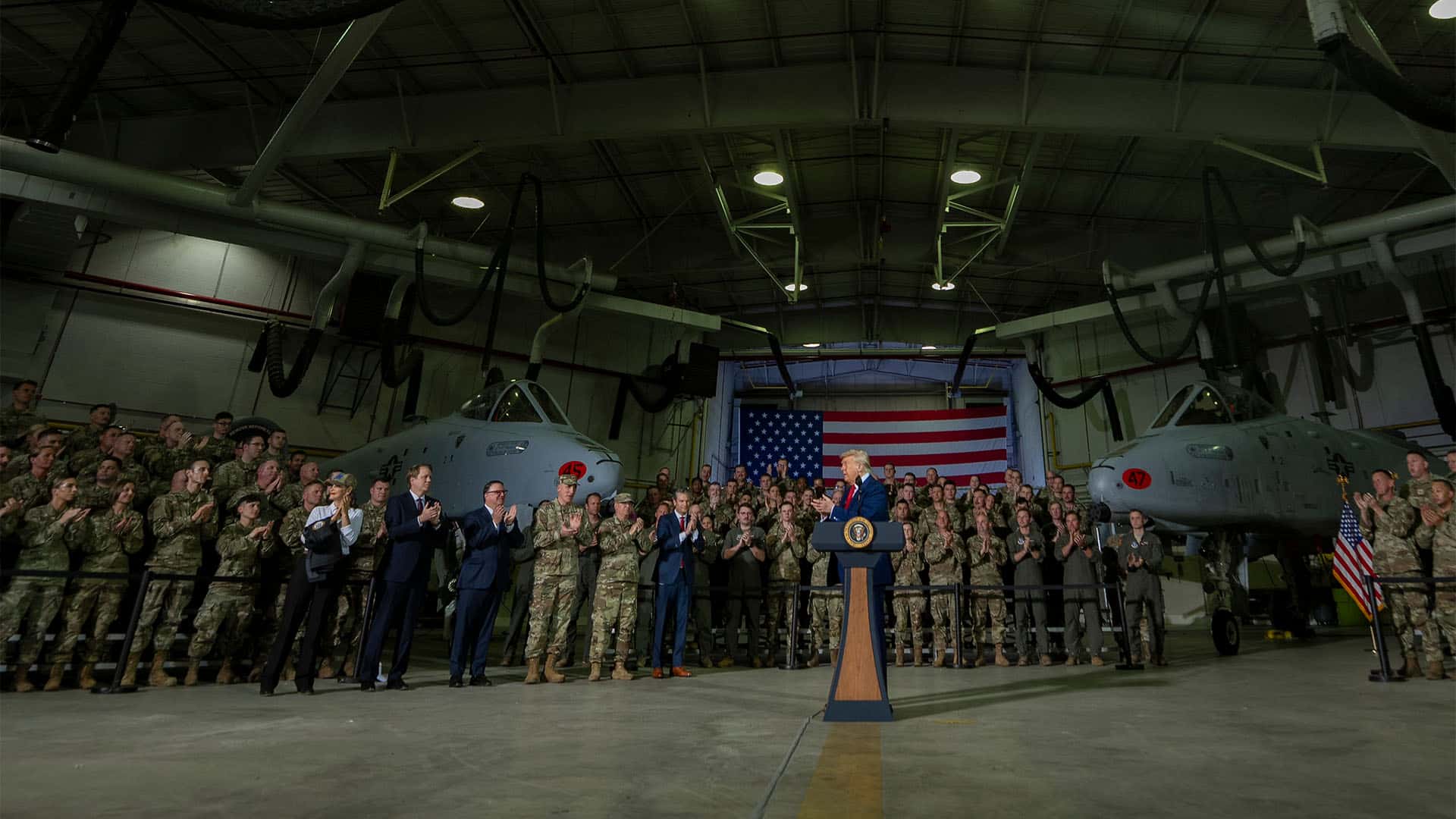Donald Trump aplaude en su discurso ante las tropas tras una tribuna con el logo del presidente (25/04/2025). A su izquierda, lo acompañan en su visita: el secretario de Defensa, Pete Hegseth y autoridades militares y políticas de Míchigan. A su alrededor, los soldados y dos aviones de la base y, al fondo, una gran bandera de Estados Unidos. Están en una nave industrial de la Base Aérea de la Guardia Nacional de Selfrifge, Míchigan.