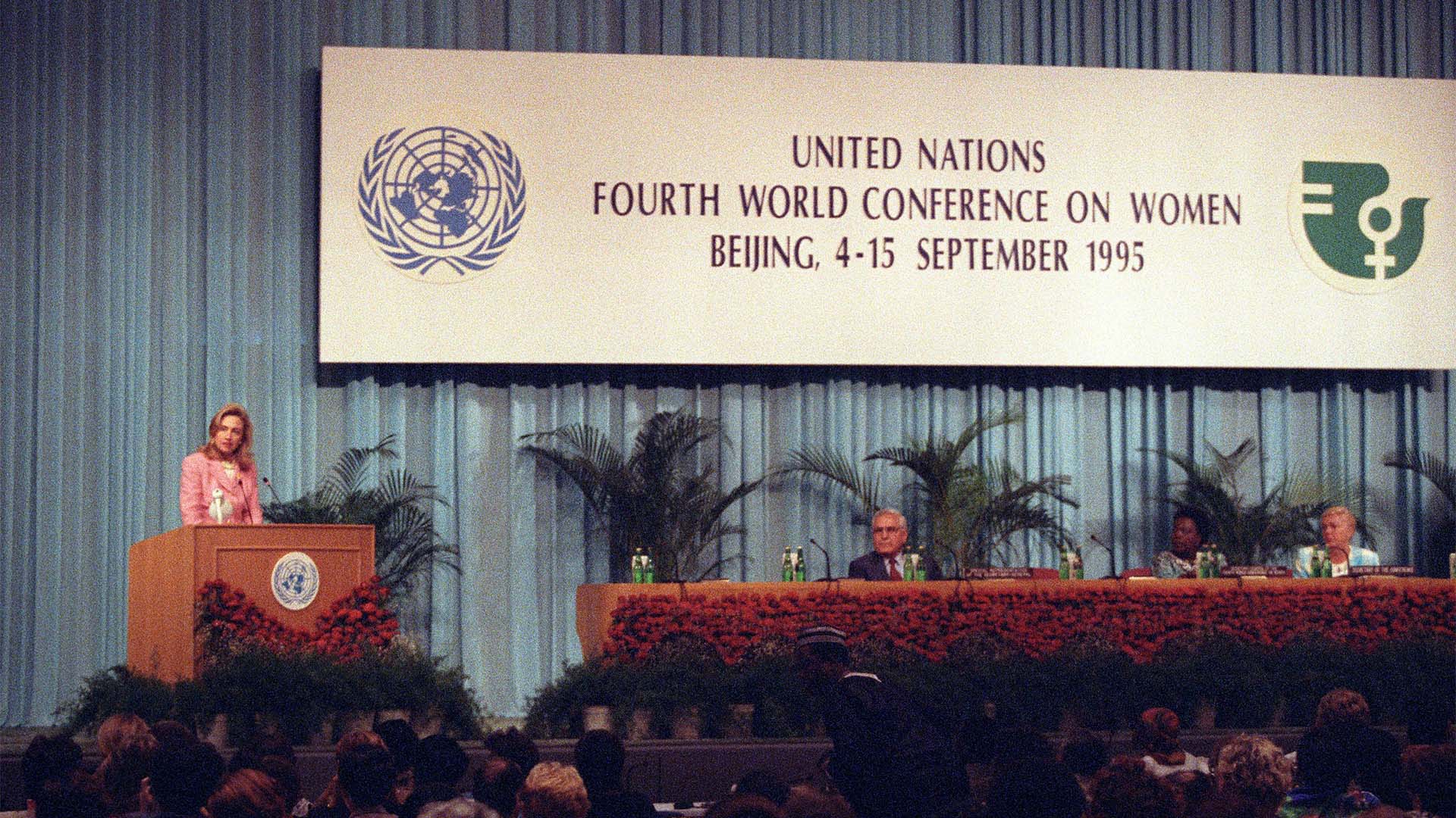 Hillary Clinton pronuncia su discurso en la IV Conferencia Mundial sobre la Mujer (Beijing, 5 de septiembre de 1995) en el escenario del Beijing International Conference Center. Viste un traje rosa y se dirige al público desde una tribuna adornada con flores y el logo de la ONU. A la derecha, se observa otra mesa con representantes y micrófonos, también decorada. Al fondo, un telón azul, un gran cartel con el texto: “United Nations Fourth World Conference on Women. Beijing, 4-15 September 1995” en la parte superior y, debajo, algunas palmeras.