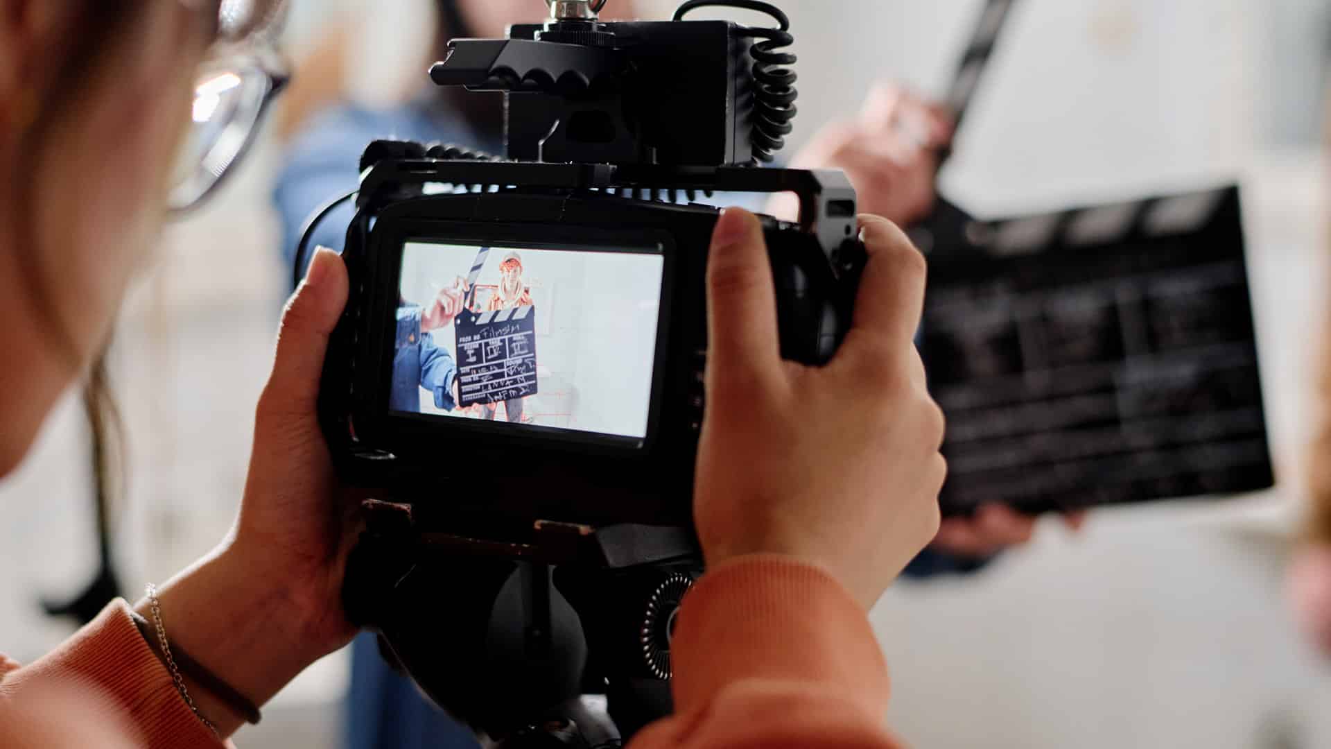 Persona sosteniendo una cámara profesional que enfoca a dos individuos frente a un fondo claro; uno sostiene una claqueta de cine con texto visible, mientras en segundo plano aparece otra claqueta desenfocada, indicando una grabación audiovisual.
