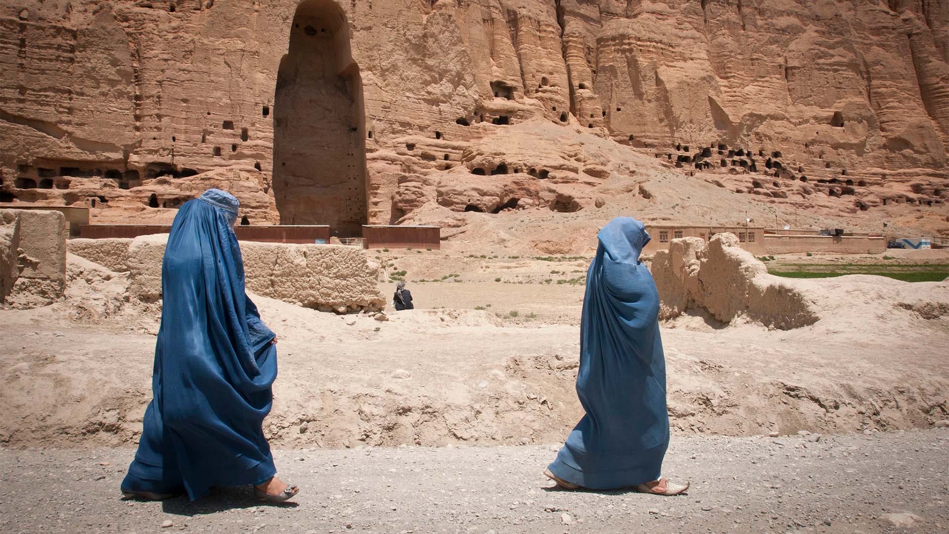 Dos mujeres caminan cubiertas con burkas de color azul en Bamiyán, provincia de Afganistán en 2012. Al fondo del paisaje árido, una cadena de montañas con una cavidad vacía donde se erguía uno de los Budas de Bamiyán construidos en los años 507 y 554 d.C. y destruidos por los talibanes en 2001.