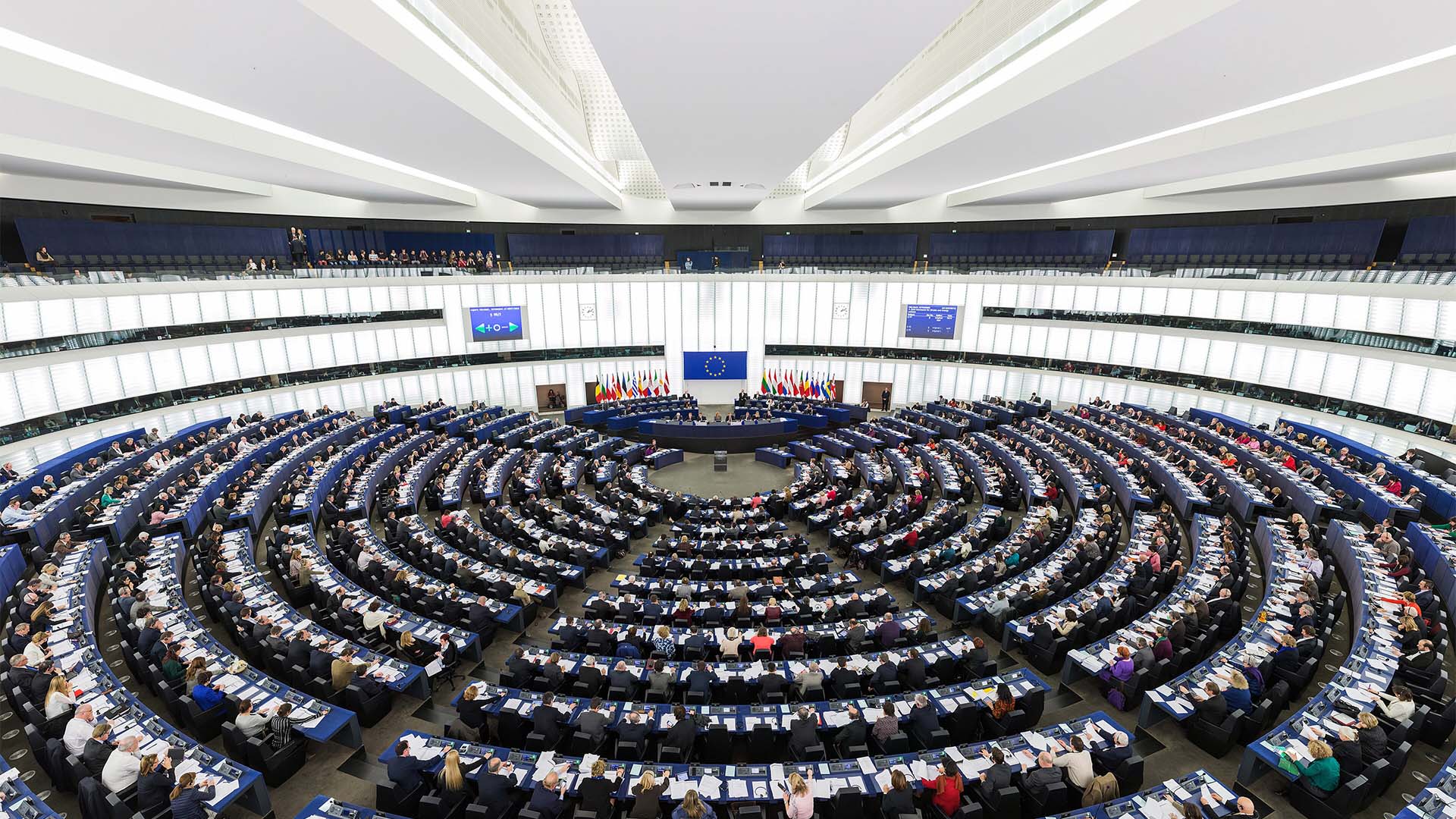 Hemiciclo del Parlamento Europeo en Estrasburgo. Siete filas de mesas con los representantes del parlamento europeo atienden a la asamblea, frente a la mesa de dirección. Detrás, pueden verse las banderas de los estados miembros y la de la Unión Europea, y dos pantallas. Tanto las paredes del hemiciclo como la cubierta, son de color blanco.