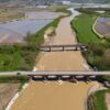 Aerial view of the riverbed after flooding from the Cervo torrent in Piedmont, Italy (27/04/2025). Among rice fields, there is a road bridge crossing the river in the foreground and a railway bridge further away, with a blue sky in the background.