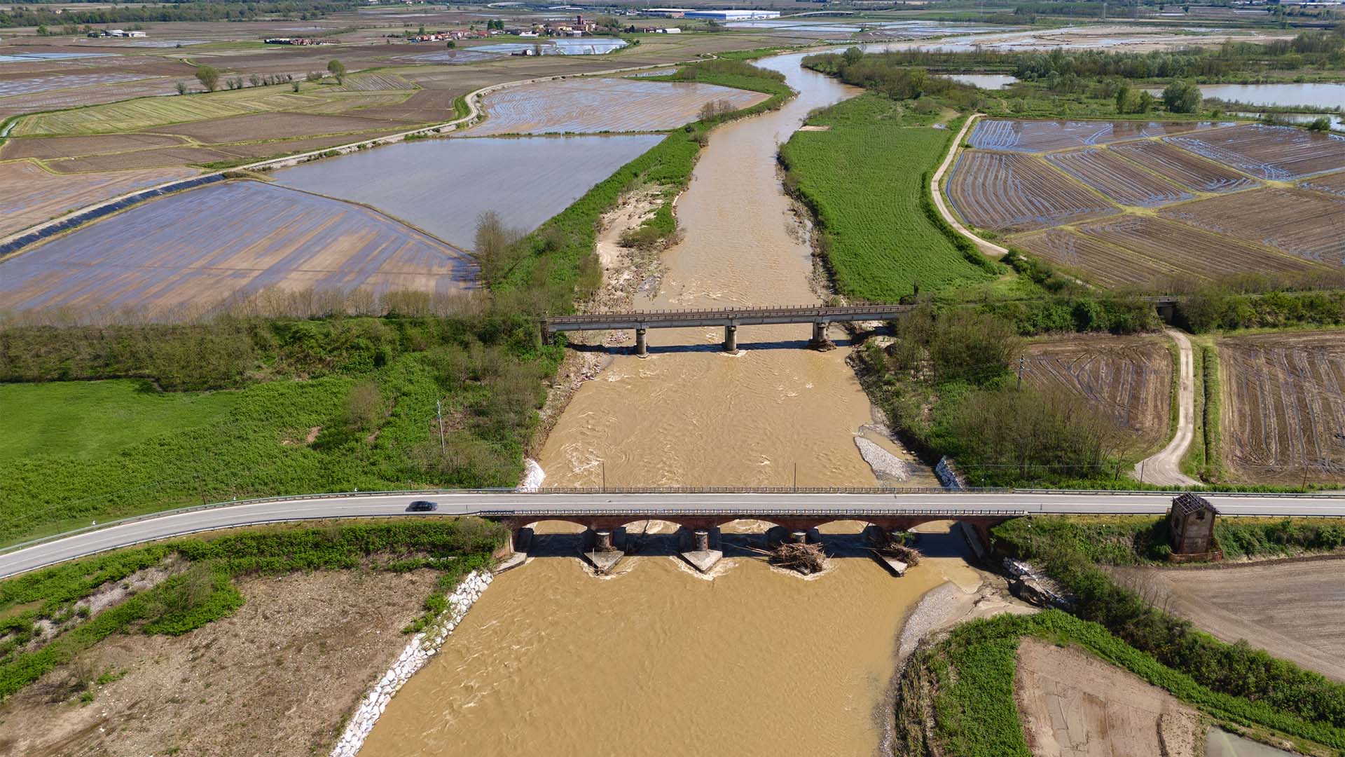 Aerial view of the riverbed after flooding from the Cervo torrent in Piedmont, Italy (27/04/2025). Among rice fields, there is a road bridge crossing the river in the foreground and a railway bridge further away, with a blue sky in the background.
