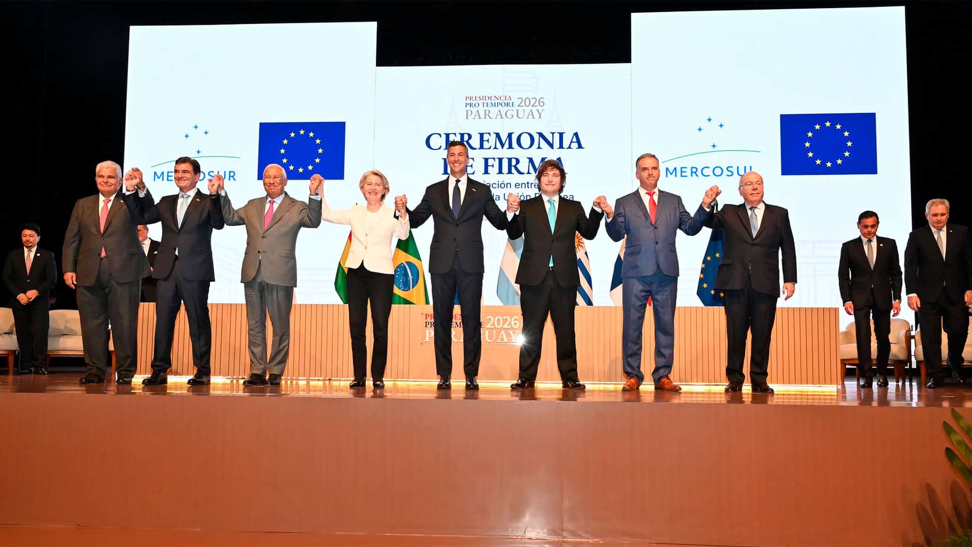 Ceremonia de la firma del acuerdo UE-Mercosur en Asunción, Paraguay (17/01/2026). Sobre el escenario, levantan las manos, unidos, de izquierda a derecha: José Raúl Mulino, presidente de Panamá; Rodrigo Paz, presidente de Bolivia; Antonio Costa, presidente del Consejo Europeo; Ursula Von der Leyen, presidenta de la Comisión Europea; Santiago Peña, presidente de Paraguay; Javier Milei, presidente de Argentina; Yamandú Orsi, presidente de Uruguay; y Mauro Vieira, ministro brasileño de Exteriores. Detrás de ellos, una mesa en la que se puede leer: “Presidencia Pro Tempore Paraguay 2026”, se ve parte de las banderas de los países firmantes y, más atrás, una pantalla: en la que se lee “Ceremonia de firma: Asociación entre el Mercosur y la Unión Europea”, enmarcado por las banderas de Mercosur y la Unión Europea.