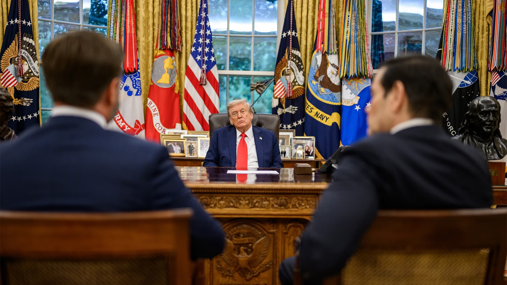 Trump, en el centro, se reúne, en el despacho oval de la Casa Blanca con el vicepresidente de EEUU, JD Vance, a la izquierda, y el secretario de Estado, Marco Rubio, a la derecha, que aparecen desenfocados. Detrás del presidente, varias fotografías, y banderas: de EEUU, la presidencia, y las fuerzas militares de Estados Unidos. Al fondo, unas ventanas con un paisaje verde difuminado. Se trata de un encuentro previo a una llamada con el presidente ruso Vladimir Putin.