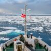 Rompehielos navegando en el mar helado en la costa norte de Groenlandia. Se puede ver la parte delantera del barco, de color blanco, de suelo verde, varias escaleras y la cabina de mando. En la proa del barco, hay varios turistas que observan el mar, y un mástil con dos banderas: una de ellas, la de Dinamarca y, la otra, aunque arrugada por el viento, la de Groenlandia.