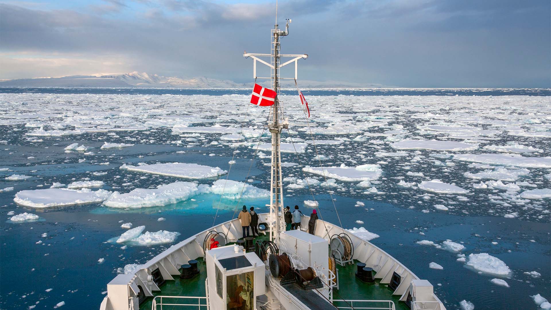 Rompehielos navegando en el mar helado en la costa norte de Groenlandia. Se puede ver la parte delantera del barco, de color blanco, de suelo verde, varias escaleras y la cabina de mando. En la proa del barco, hay varios turistas que observan el mar, y un mástil con dos banderas: una de ellas, la de Dinamarca y, la otra, aunque arrugada por el viento, la de Groenlandia.