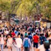 Crowds of people walking along La Rambla in Barcelona, surrounded by stalls and shops on both sides of the street, with tall trees providing shade overhead.