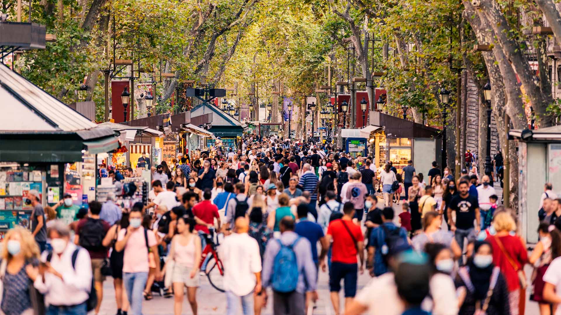 Crowds of people walking along La Rambla in Barcelona, surrounded by stalls and shops on both sides of the street, with tall trees providing shade overhead.
