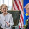 Ursula Von der Leyen y Donald Trump en la firma del acuerdo comercial bilateral (27/07/2025) en el Trump Turnberry golf course, en Turnberry, Escocia. Von der Leyen, sentada en una butaca verde, a la izquierda, habla y enfatiza con sus manos, a Donald Trump, desenfocado, a la derecha. Al fondo, pueden verse, desenfocadas, la bandera de EEUU y la de la Unión Europea, y, parte de la sala donde ambos representantes firman el acuerdo bilateral.