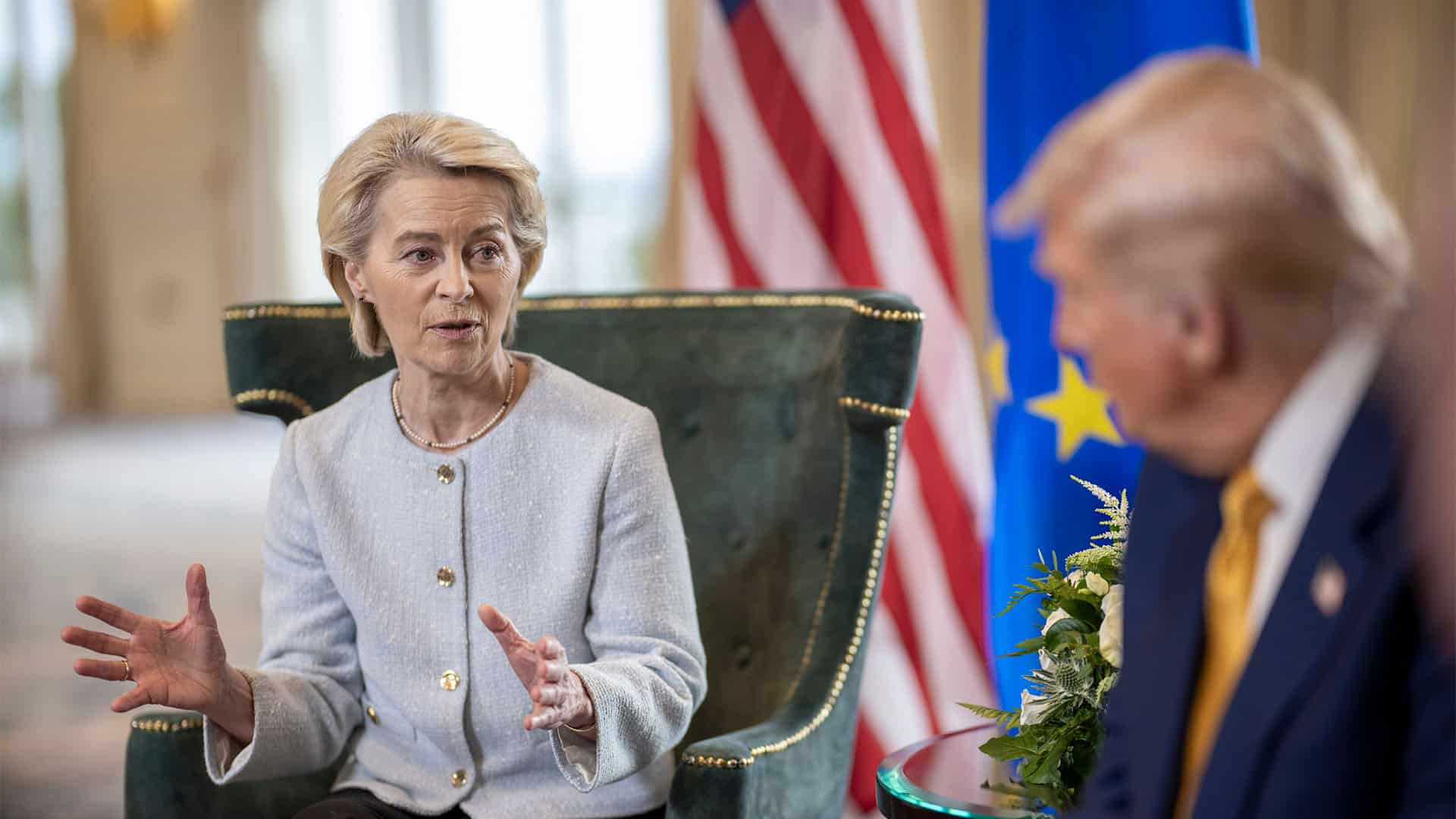 Ursula Von der Leyen y Donald Trump en la firma del acuerdo comercial bilateral (27/07/2025) en el Trump Turnberry golf course, en Turnberry, Escocia. Von der Leyen, sentada en una butaca verde, a la izquierda, habla y enfatiza con sus manos, a Donald Trump, desenfocado, a la derecha. Al fondo, pueden verse, desenfocadas, la bandera de EEUU y la de la Unión Europea, y, parte de la sala donde ambos representantes firman el acuerdo bilateral.