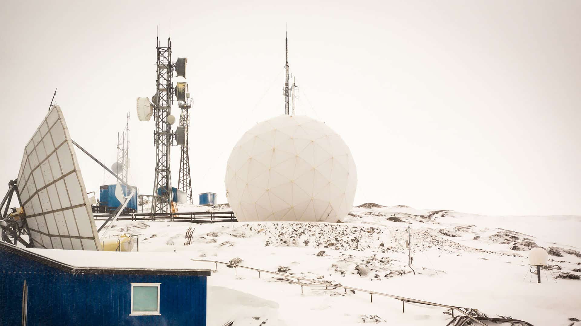 Estación de telecomunicaciones en Nuuk, Groenlandia. Sobre las rocas nevadas, se puede observar la estación de comunicación, con varias antenas, torres y casetas de color azul, que contrastan con el resto de los elementos, predominantemente blancos.