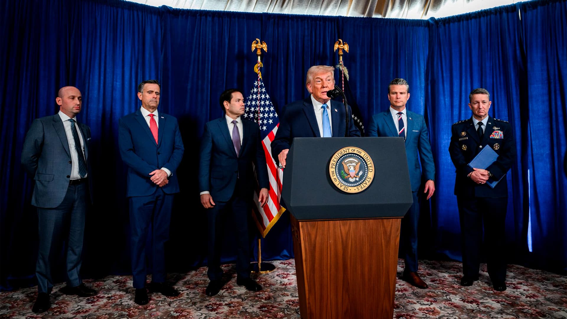 Donald Trump en la rueda de prensa tras la “Operación Resolución Absoluta” en Venezuela y la captura de Nicolás Maduro (3 de enero de 2026) en Mar-a-Lago, Florida. El presidente habla tras el atril, adornado con el logo del presidente y un micrófono. Detrás de él, de izquierda a derecha, Stephen Miller, asesor de Seguridad Nacional; John Ratcliffe, director de la CIA; Marco Rubio, secretario de Estado; Pete Hegseth, secretario de Guerra; y el general Dan Caine, jefe del Estado Mayor Conjunto. Detrás, las banderas de EEUU y la presidencia, y un telón azul y blanco.