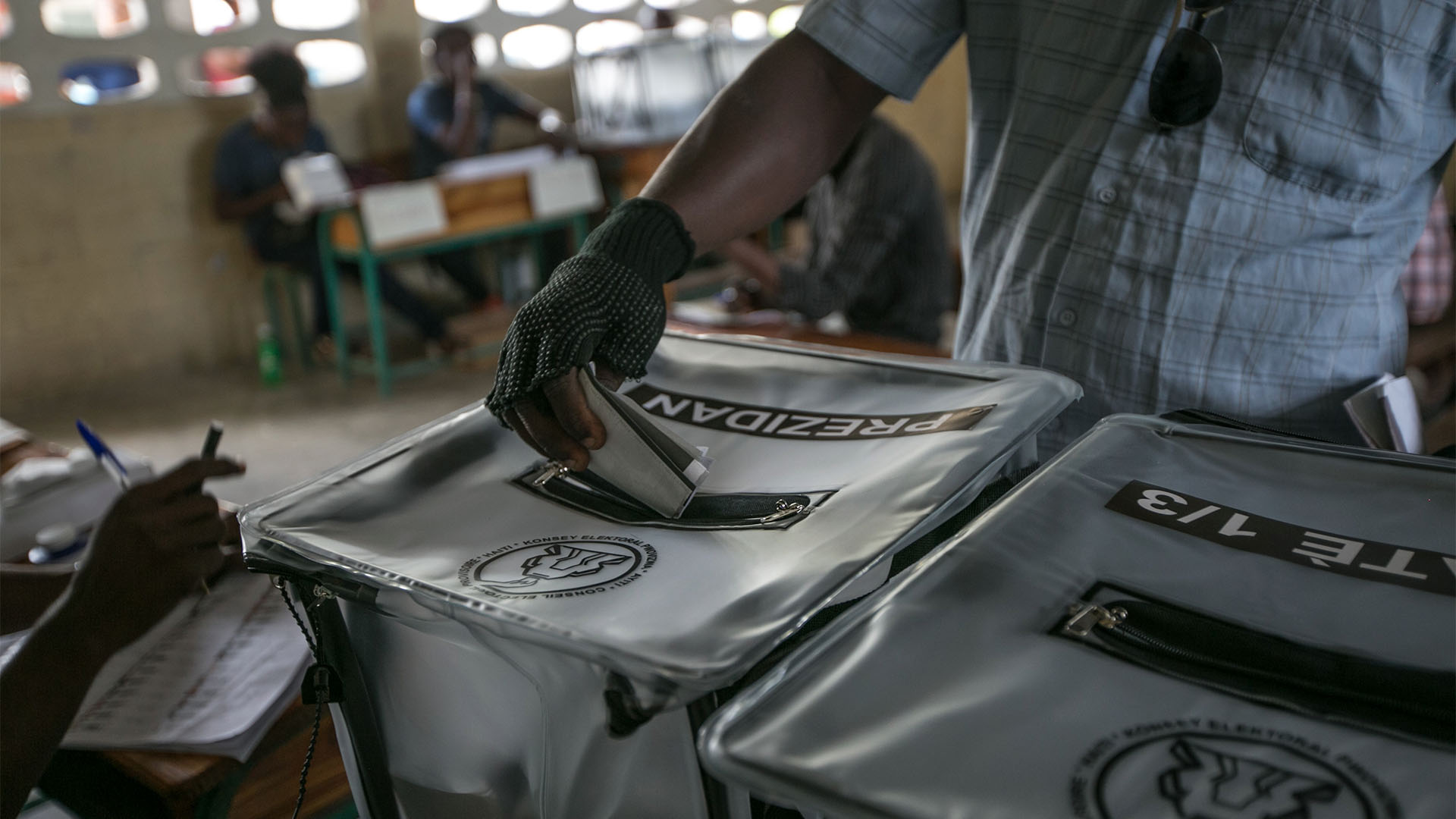 Una persona introduce la papeleta de voto para las elecciones presidenciales de Haití, en Puerto príncipe (20/11/2016). La urna donde introduce el voto es de plástico opaco, donde puede leerse la palabra “Prezidan”. A la izquierda, puede verse otra persona registrando los votos, y, al fondo, desenfocada, otra mesa electoral, con urnas y otras dos personas gestionando los votos.