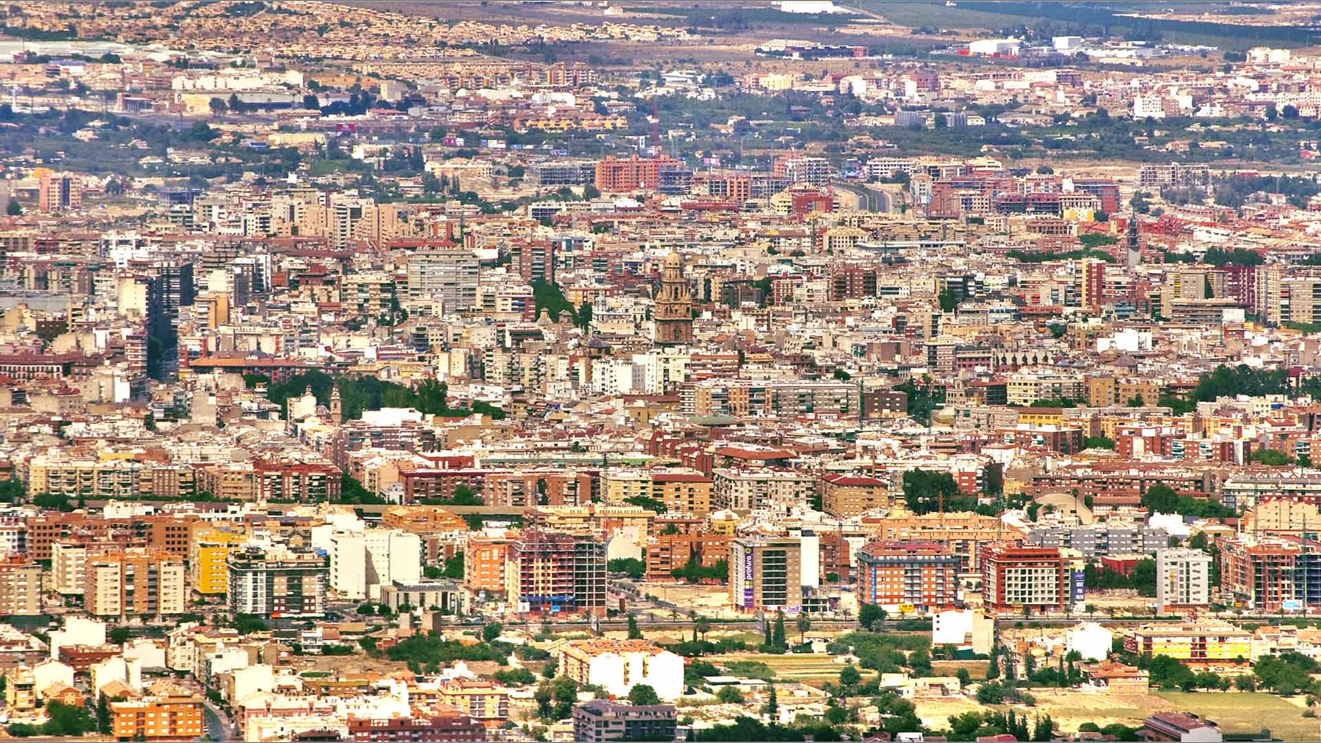 Vista aérea de la ciudad de Murcia. Cientos de edificios de diferentes alturas y colores, donde destaca la torre de la Catedral de la ciudad, en el centro, de color amarronado. Al fondo, algunas concentraciones de árboles y vegetación, aunque el paisaje es árido.