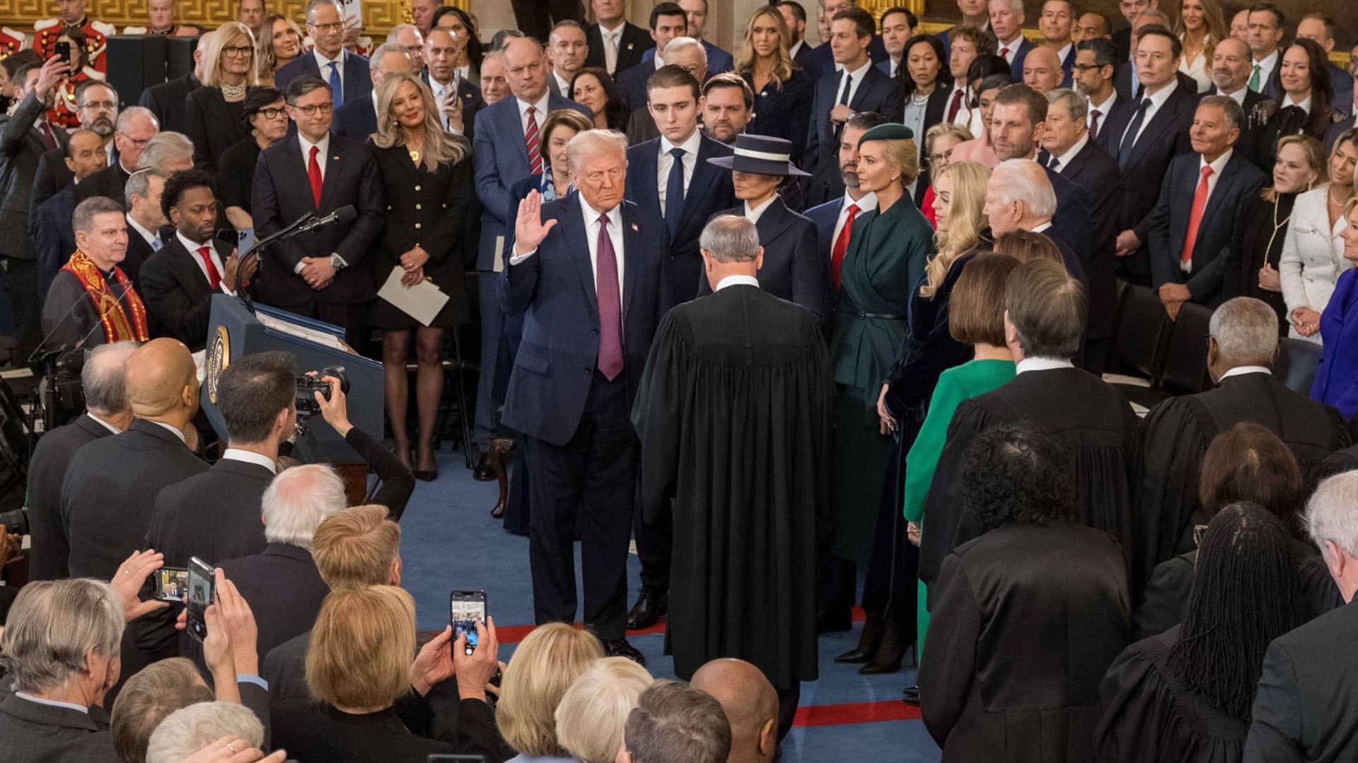 Donald Trump, con la mano levantada, presta juramento al cargo en su segundo mandato como presidente de EEUU ante el presidente del Tribunal Supremo, John Roberts, en la Rotonda del Capitolio (20/01/2025). Lo rodean personalidades políticas y empresariales destacadas, que fotografían la escena con cámaras y teléfonos móviles.
