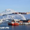 Vista del puerto Adolf Jensensvej en Nuuk (Groenlandia), con un paisaje ártico con barcos rojos atracados, edificios coloridos en la costa y la montaña Sermitsiaq nevada al fondo bajo un cielo despejado.