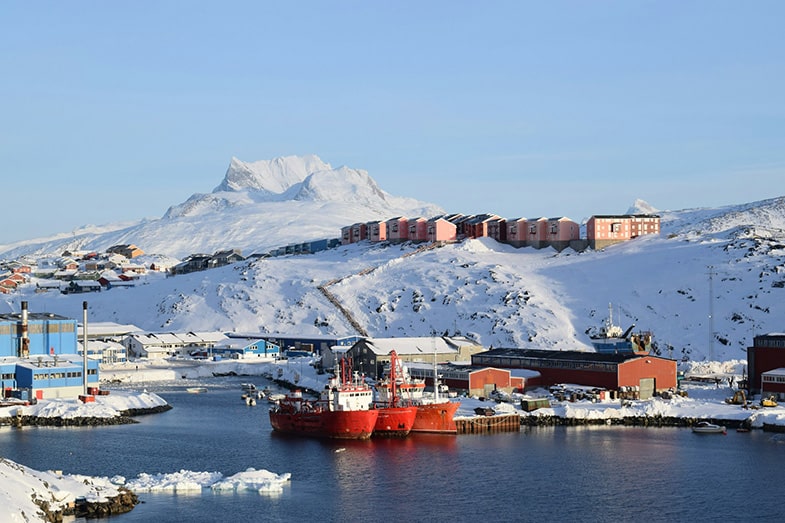 Vista del puerto Adolf Jensensvej en Nuuk (Groenlandia), con un paisaje ártico con barcos rojos atracados, edificios coloridos en la costa y la montaña Sermitsiaq nevada al fondo bajo un cielo despejado.
