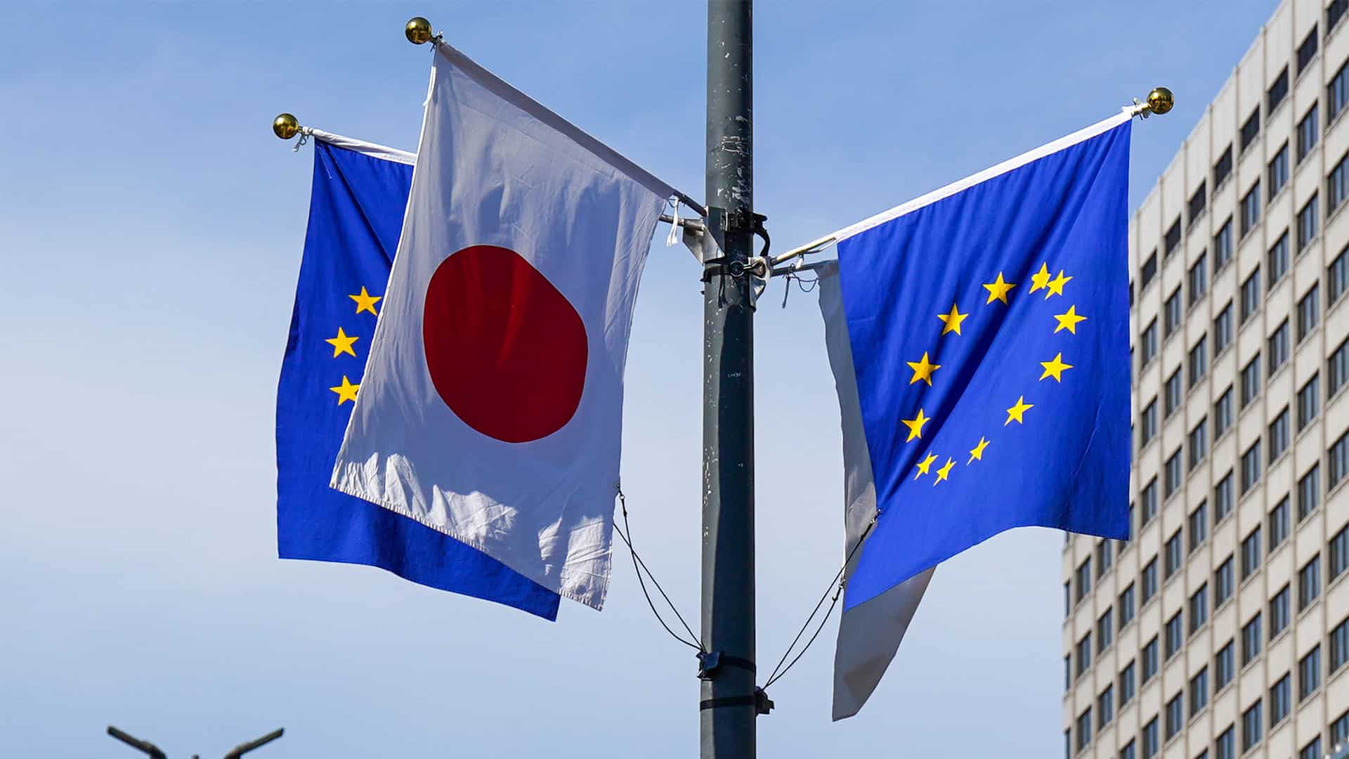 Mástil con las banderas de la Unión Europea y Japón, durante la visita de Kaja Jallas a Japón en julio de 2025. El mástil tiene cuatro postes con cuatro banderas, dos de la UE y dos de Japón. Detrás de las banderas, un edificio blanco con numerosas ventanas y un cielo completamente azul y despejado.