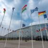 Low-angle view of the 32 flags of NATO member countries flying in a circle. In the background, a NATO flag can be seen, and further behind, the glass building of its headquarters in Brussels.