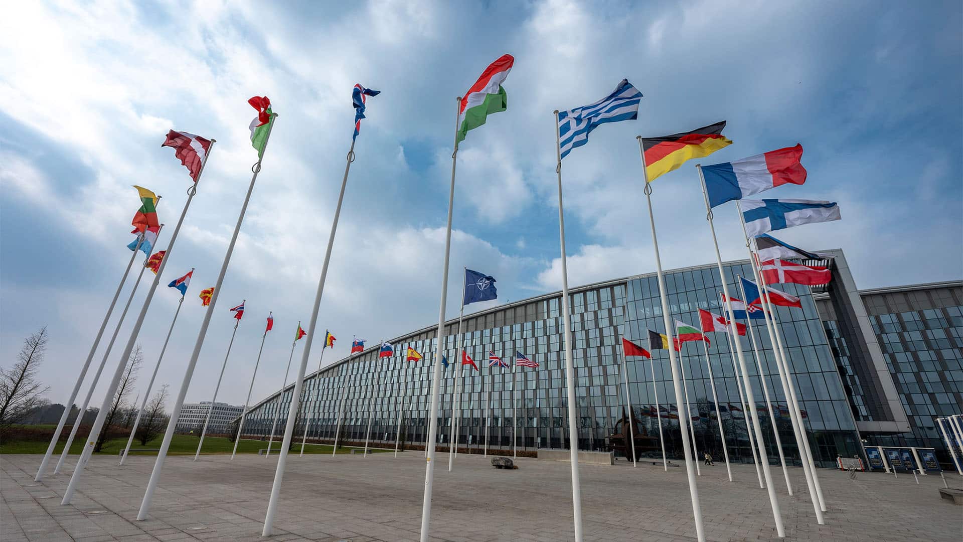 Low-angle view of the 32 flags of NATO member countries flying in a circle. In the background, a NATO flag can be seen, and further behind, the glass building of its headquarters in Brussels.
