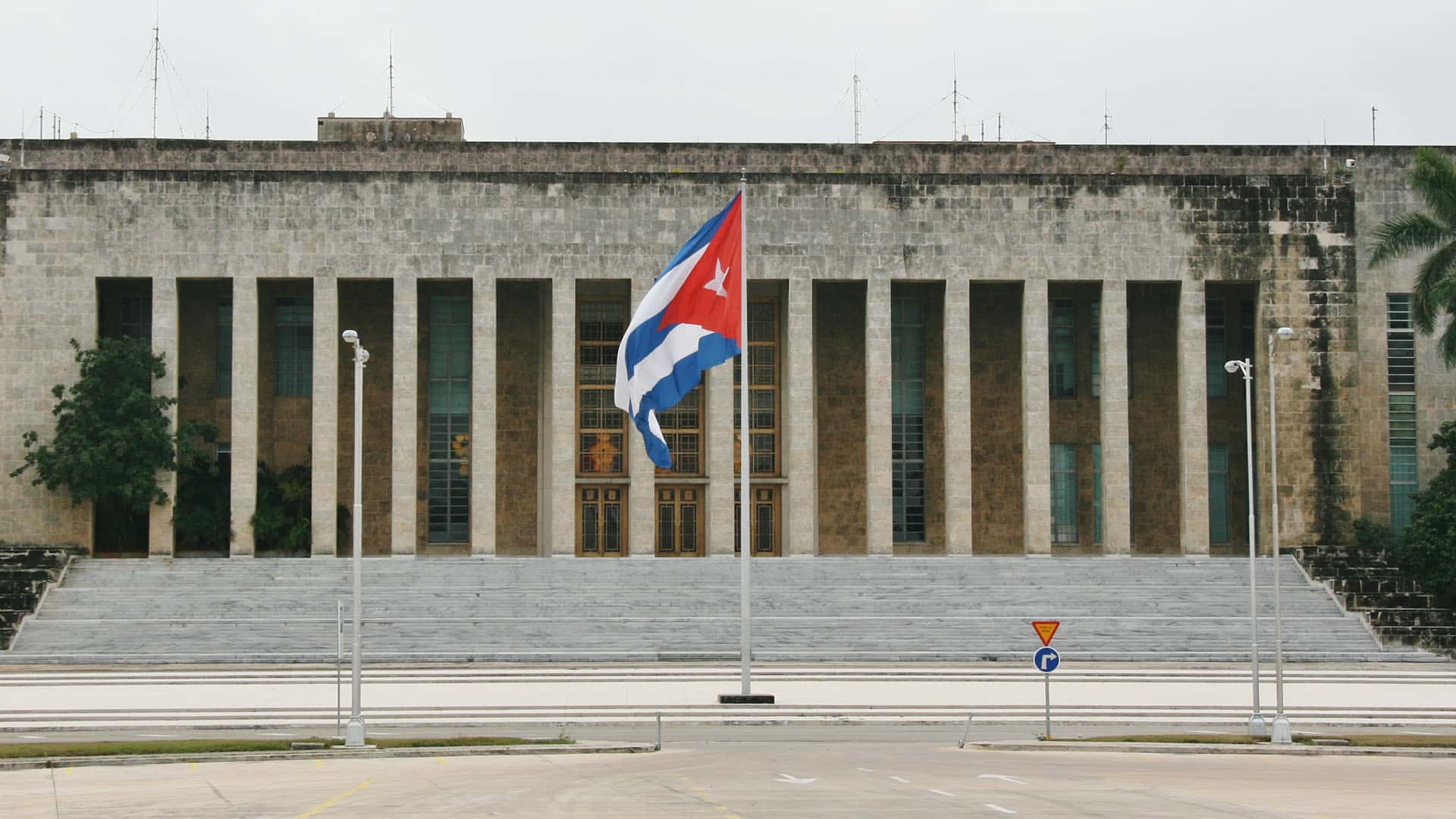 Vista de la fachada principal del Palacio de la Revolución, sede de la presidencia y gobierno de la República de Cuba. La fachada cuenta con múltiples columnas tras la escalinata principal de entrada. En el centro de la imagen, la bandera de Cuba ondea en un mástil.