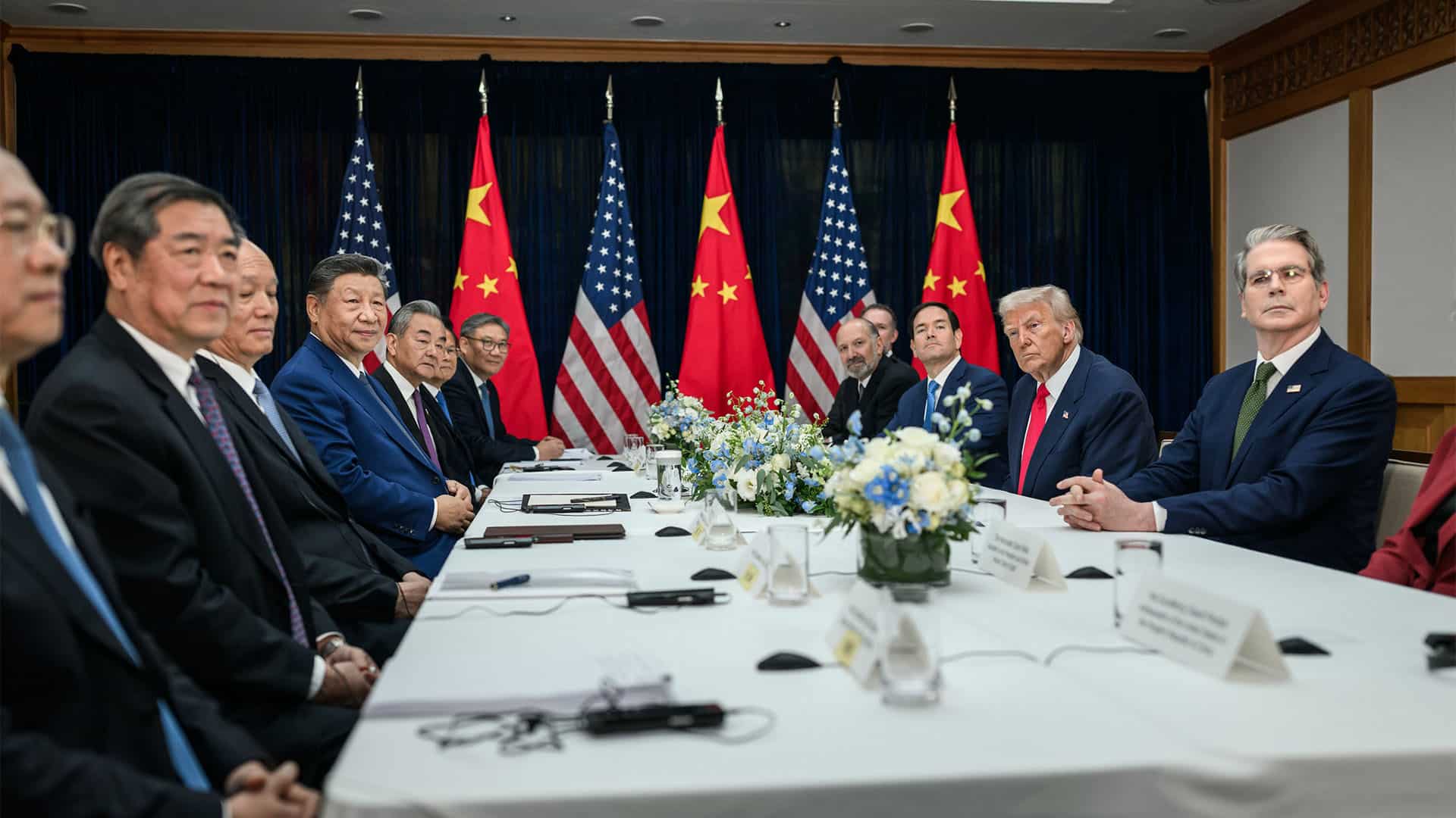 Reunión bilateral entre Trump y Xi Jinping (30/10/2025) en el aeropuerto internacional de Gimhae, en Busán, Corea del Sur. A la izquierda, los representantes de la administración de China, liderada por Xi Jinping; y, a la derecha, la de EEUU, encabezada por Trump, miran hacia la cámara que toma la fotografía. Al fondo, tres banderas de EEUU y tres de China, intercaladas.