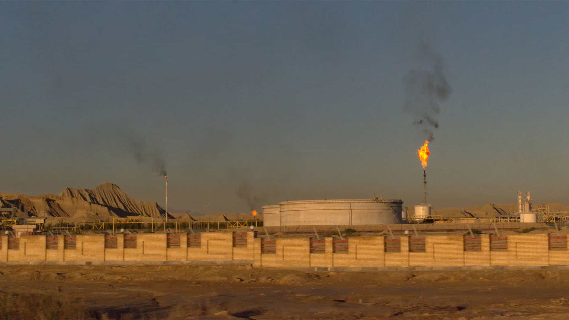 Una planta de refinería de petróleo y una empresa de gas natural en el Golfo Pérsico, Irán. La refinería, de color blanco y delimitada por un muro de ladrillos también blanco, tiene varias llamas que humean en el aire. Al fondo, un paisaje montañoso y un cielo turbio.