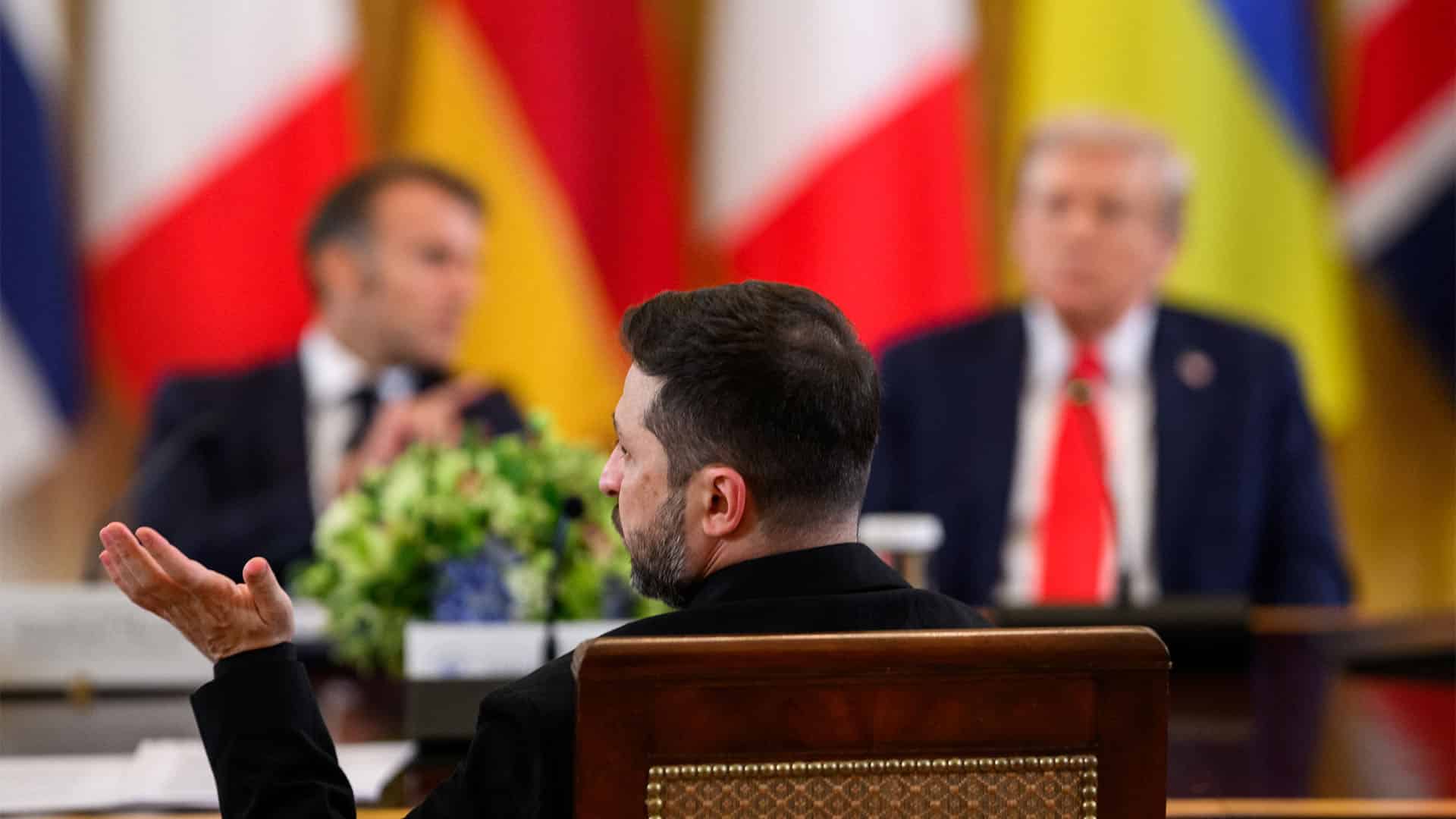 En primer plano, Volodímir Zelenski, presidente de Ucrania, enfatiza con su mano mientras habla. Detrás de él, desenfocados, Emmanuel Macron, presidente de la República Francesa, a la izquierda, y Donald Trump, presidente de EEUU, a la derecha. Los representantes se reúnen en la Casa Blanca, en un encuentro multilateral organizado por el presidente Trump con los líderes europeos.