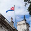 Contrapicado de la bandera de Cuba ondeando sobre un cielo claro. El mástil está colocado en la parte superior de la fachada de piedra del Palacio de los Capitanes Generales. A su derecha, se eleva un muro más alto con una campana.