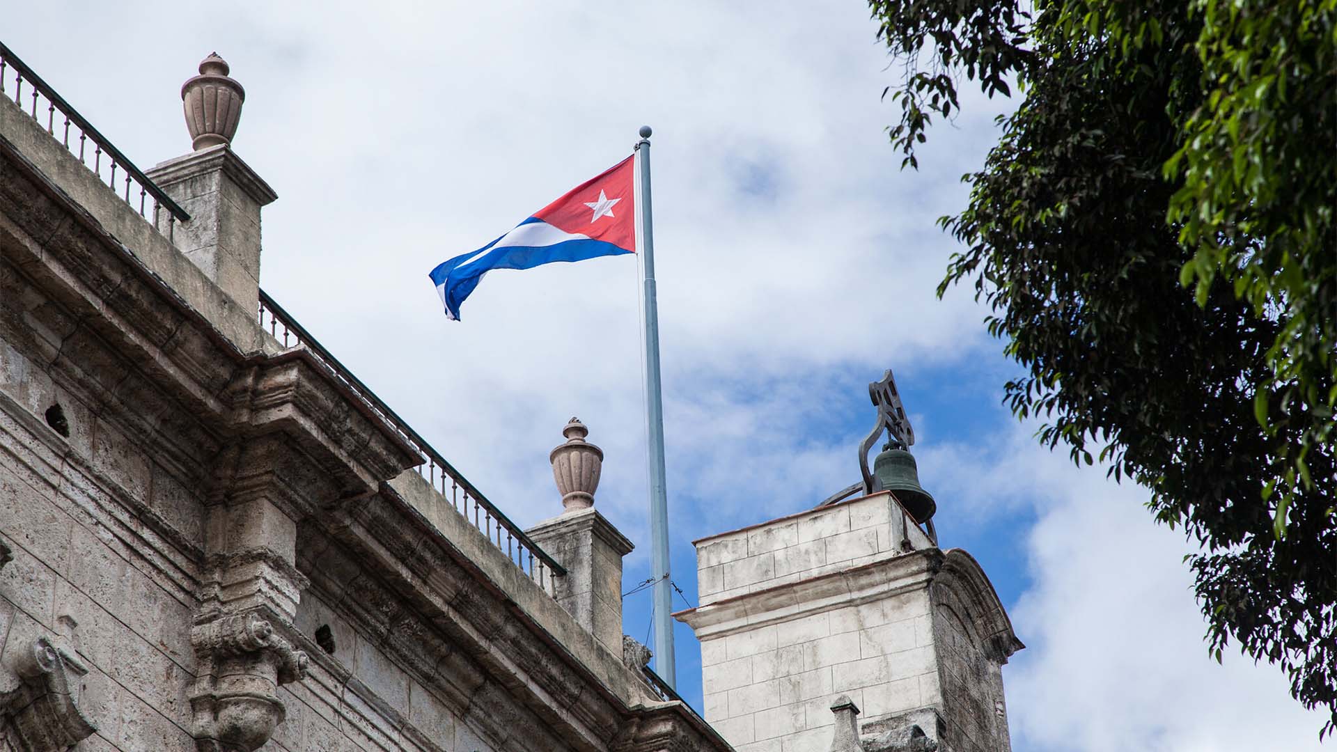 Contrapicado de la bandera de Cuba ondeando sobre un cielo claro. El mástil está colocado en la parte superior de la fachada de piedra del Palacio de los Capitanes Generales. A su derecha, se eleva un muro más alto con una campana.
